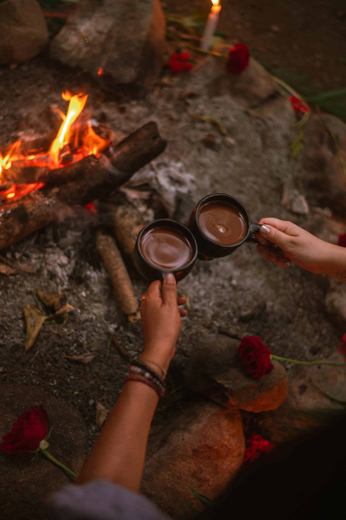 Two cups of cacao held by hands near a fire, rose petals